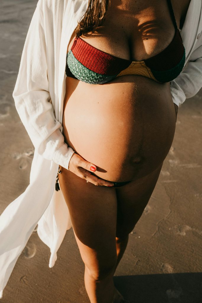 A pregnant woman in a bikini holding her belly on a sunny beach.